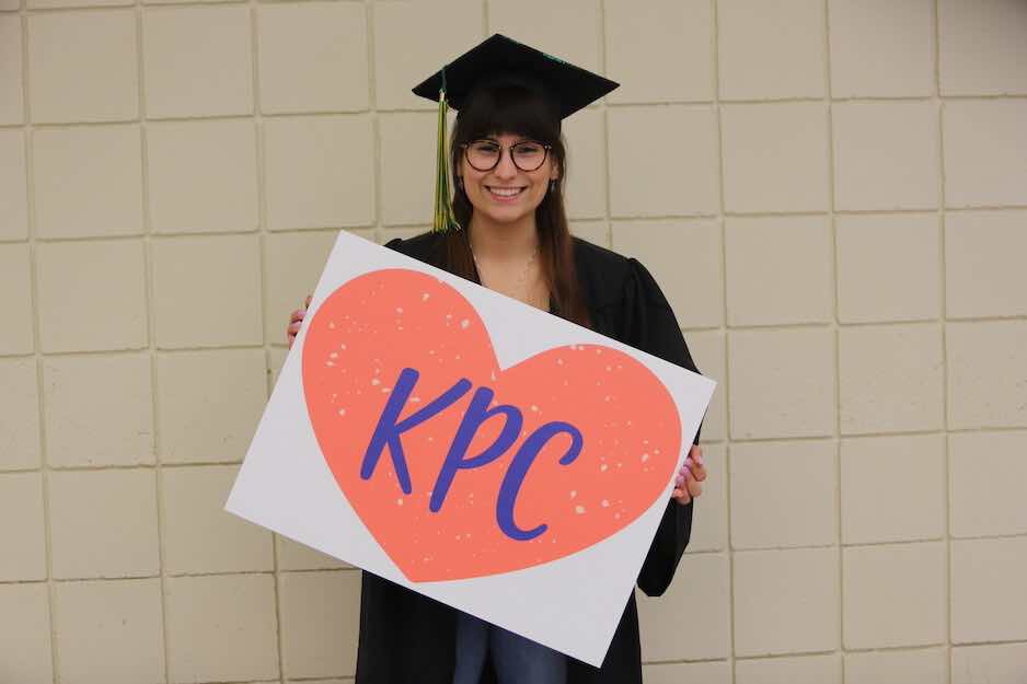 Girl with classes wearing graduation cap and gown holding a sign of a red heart that says KPC